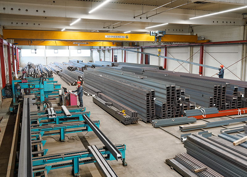 Innenansicht einer großen Lagerhalle, in der Formstahl in unterschiedlichen Profilformen, Größen und Längen ordentlich gestapelt liegt. Ein gelbes Brückenkransystem verläuft an der Hallendecke und eine Säge steht bereit für den Zuschnitt der Stahlträger. Die Halle ist entsprechend strukturiert und für den kurzfristigen Materialumschlag ausgelegt.