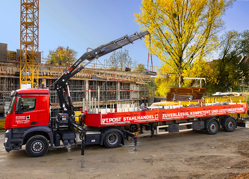 Ein LKW mit integriertem Entladekran liefert gerade Stahl auf einer Großbaustelle an. Der Kran ist im Einsatz und hebt die schweren Stahlteile vom LKW auf die Baustellenfläche. Im Hintergrund sind Baugerüste und Rohbauten sichtbar.